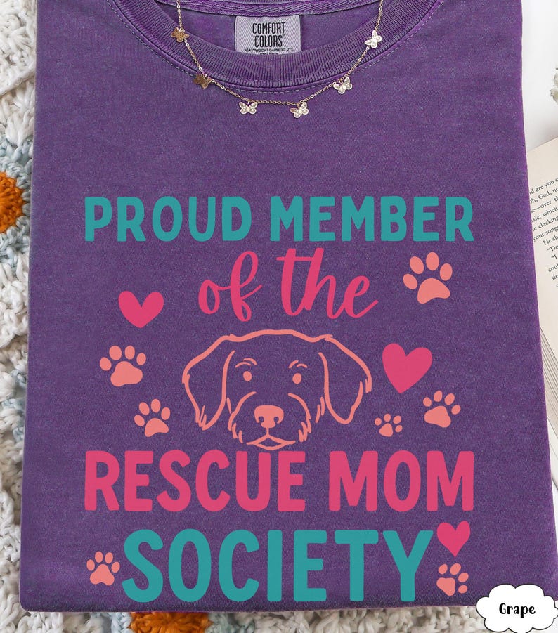 May include: A purple Comfort Colors t-shirt with the text "PROUD MEMBER of the RESCUE MOM SOCIETY" in teal and pink. The design includes paw prints, hearts, and a dog illustration. A gold butterfly necklace is draped on the shirt.