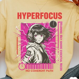 May include: Yellow t-shirt featuring a graphic of an anime-style character wearing headphones, set against a pink background with the text "HYPERFOCUS" and "NO COHERENT PATH". The design includes a clock and the warning "MAY IGNORE ESSENTIAL NEEDS".