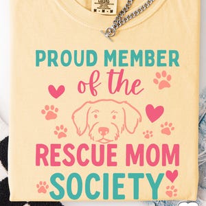 May include: A light yellow t-shirt with the text "PROUD MEMBER of the RESCUE MOM SOCIETY" in teal and pink. The design includes paw prints, hearts, and a dog illustration. A silver chain is visible at the top.
