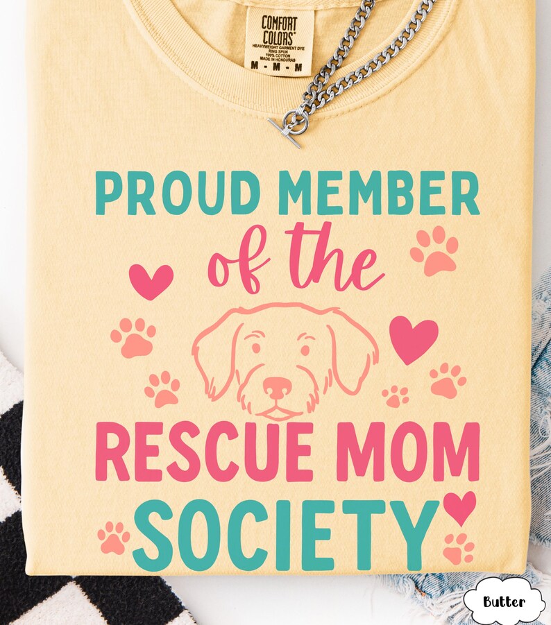 May include: A light yellow t-shirt with the text "PROUD MEMBER of the RESCUE MOM SOCIETY" in teal and pink. The design includes paw prints, hearts, and a dog illustration. A silver chain is visible at the top.