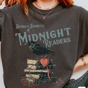 May include: Dark gray t-shirt with the text "Secret Society of Midnight Readers." A black crow sits atop a stack of books tied with a red heart and ribbon. The shirt is a comfortable, casual style.