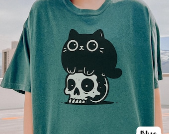 May include: Blue Spruce t-shirt featuring a black cat sitting on a skull graphic. The cat has large eyes and a simple design. The skull is white with black details. The shirt is a solid teal color. The text "Blue Spruce" is in a white box.
