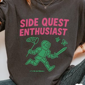 May include: Dark gray t-shirt with the text "SIDE QUEST ENTHUSIAST" in pink. A green cartoon knight with a sword and a net is running towards two green butterflies. The shirt is a casual wear item.