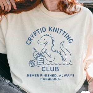 May include: Ivory sweatshirt featuring a blue dinosaur knitting graphic. The text above reads "CRYPTID KNITTING CLUB" and below "NEVER FINISHED, ALWAYS FABULOUS". The design includes a ball of yarn and knitting needles. A fun, quirky design.