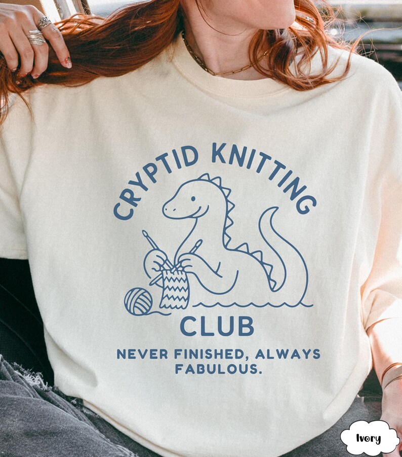 May include: Ivory sweatshirt featuring a blue dinosaur knitting graphic. The text above reads "CRYPTID KNITTING CLUB" and below "NEVER FINISHED, ALWAYS FABULOUS". The design includes a ball of yarn and knitting needles. A fun, quirky design.