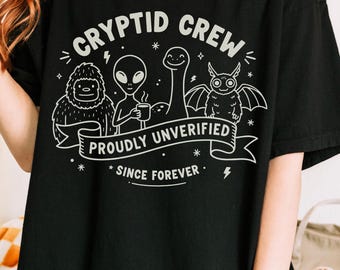 May include: Black t-shirt with a white graphic featuring the text "CRYPTID CREW" and illustrations of a Bigfoot, alien, Loch Ness Monster, and a bat-like creature. The phrase "PROUDLY UNVERIFIED SINCE FOREVER" is also included.