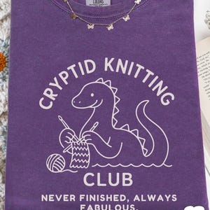 May include: A purple t-shirt featuring the words "CRYPTID KNITTING CLUB" and a cartoon dinosaur knitting. The shirt also has the text "NEVER FINISHED, ALWAYS FABULOUS." A gold butterfly necklace is draped on the shirt.