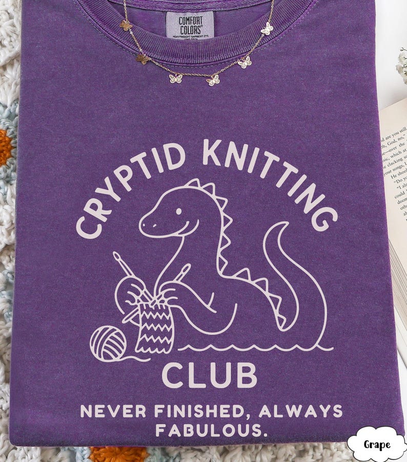 May include: A purple t-shirt featuring the words "CRYPTID KNITTING CLUB" and a cartoon dinosaur knitting. The shirt also has the text "NEVER FINISHED, ALWAYS FABULOUS." A gold butterfly necklace is draped on the shirt.