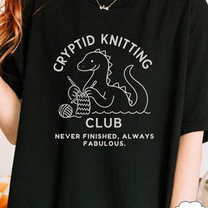 May include: Black t-shirt featuring a white graphic of a dinosaur knitting. The text says "Cryptid Knitting Club" and "Never Finished, Always Fabulous". A casual, comfortable style of shirt.