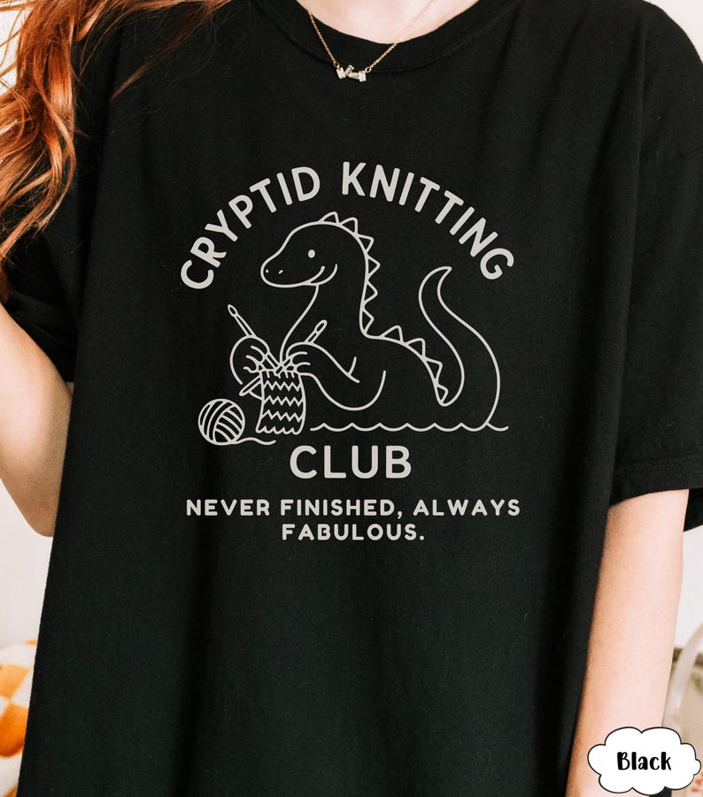 May include: Black t-shirt featuring a white graphic of a dinosaur knitting. The text says "Cryptid Knitting Club" and "Never Finished, Always Fabulous". A casual, comfortable style of shirt.