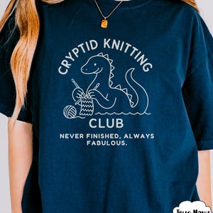 May include: Navy blue t-shirt featuring a white dinosaur knitting graphic. The text on the shirt says "Cryptid Knitting Club" and "Never Finished, Always Fabulous". The t-shirt has a relaxed fit and is a dark navy colour.