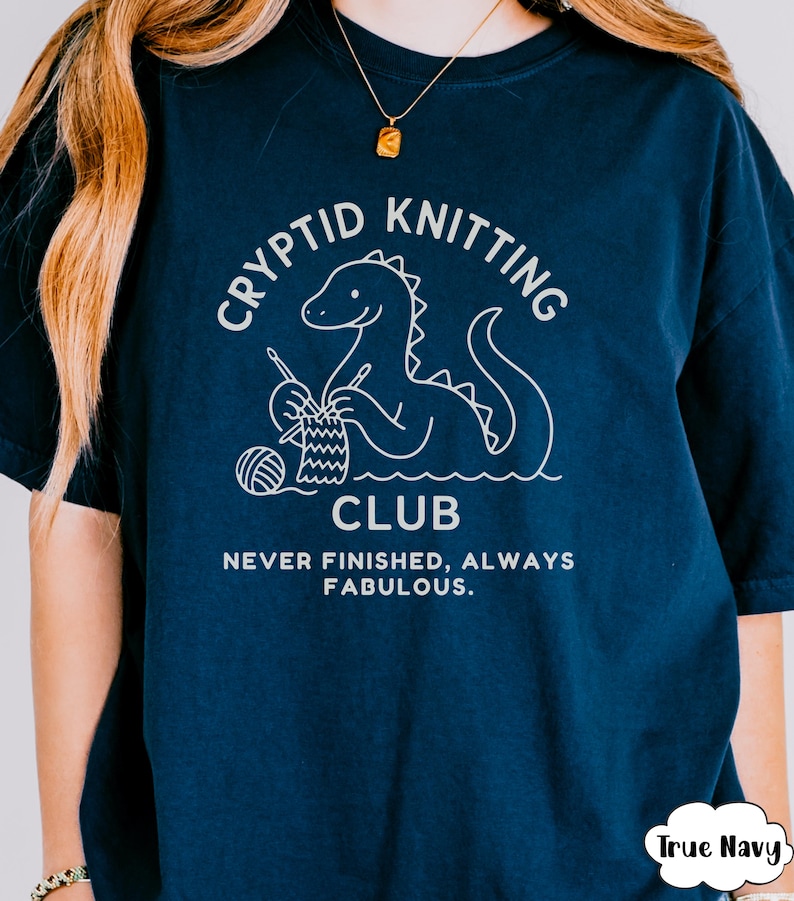 May include: Navy blue t-shirt featuring a white dinosaur knitting graphic. The text on the shirt says "Cryptid Knitting Club" and "Never Finished, Always Fabulous". The t-shirt has a relaxed fit and is a dark navy colour.
