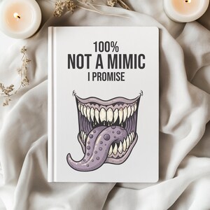 May include: A white notebook with the text "100% NOT A MIMIC I PROMISE" and a cartoon monster mouth illustration. The mouth is purple and has sharp teeth and a long tongue. The notebook is on a white cloth.