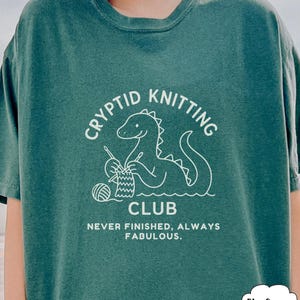 May include: Blue Spruce t-shirt featuring a white dinosaur knitting graphic. The text says "CRYPTID KNITTING CLUB" and "NEVER FINISHED, ALWAYS FABULOUS". The shirt has a relaxed fit and is made of a soft material.