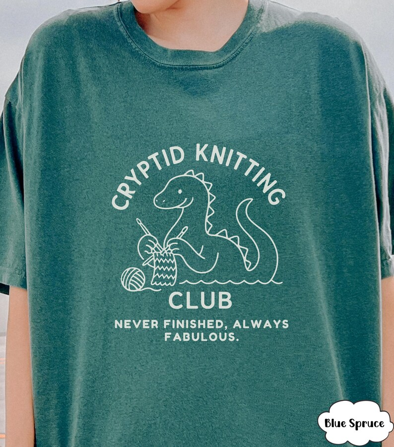 May include: Blue Spruce t-shirt featuring a white dinosaur knitting graphic. The text says "CRYPTID KNITTING CLUB" and "NEVER FINISHED, ALWAYS FABULOUS". The shirt has a relaxed fit and is made of a soft material.