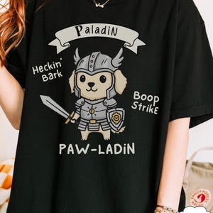 May include: Black t-shirt featuring a cartoon dog in knight's armor, holding a sword and shield. The design includes the words "Paladin," "Heckin' Bark," "Boop Strike," and "Paw-Ladin."