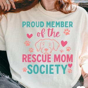 May include: Ivory-colored sweatshirt with the text "PROUD MEMBER of the RESCUE MOM SOCIETY" in teal and pink. The design includes paw prints, hearts, and a dog illustration.