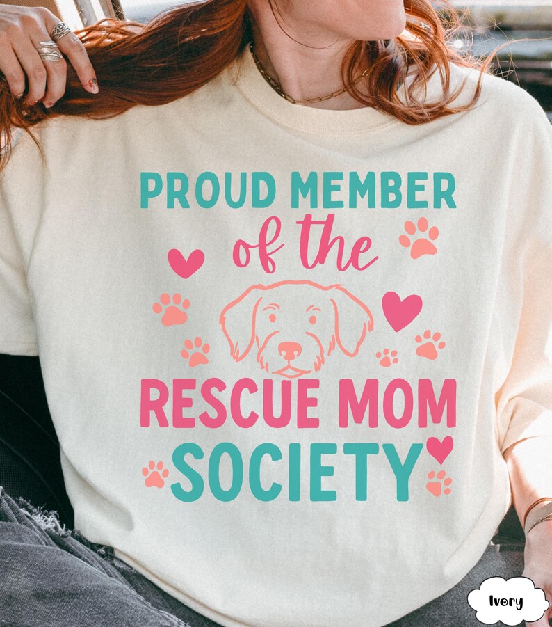May include: Ivory-colored sweatshirt with the text "PROUD MEMBER of the RESCUE MOM SOCIETY" in teal and pink. The design includes paw prints, hearts, and a dog illustration.