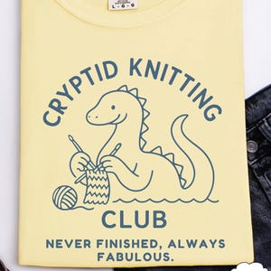 May include: A pale yellow t-shirt featuring a blue graphic of a Loch Ness Monster knitting, with the words "CRYPTID KNITTING CLUB". Below the graphic is the text "NEVER FINISHED, ALWAYS FABULOUS". The shirt is a Comfort Colors brand.