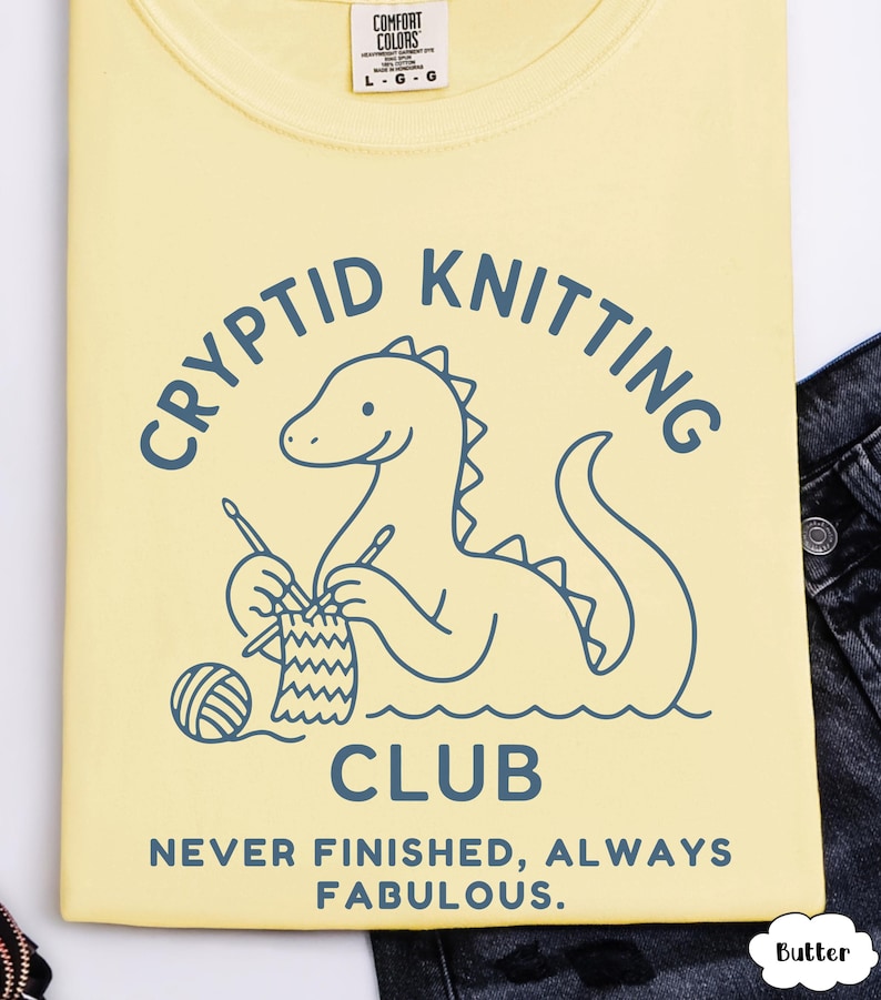 May include: A pale yellow t-shirt featuring a blue graphic of a Loch Ness Monster knitting, with the words "CRYPTID KNITTING CLUB". Below the graphic is the text "NEVER FINISHED, ALWAYS FABULOUS". The shirt is a Comfort Colors brand.