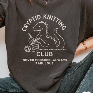 May include: Charcoal grey t-shirt featuring a white illustration of a dinosaur knitting. The text reads "Cryptid Knitting Club" above the image and "Never Finished, Always Fabulous" below. The graphic includes yarn and needles.