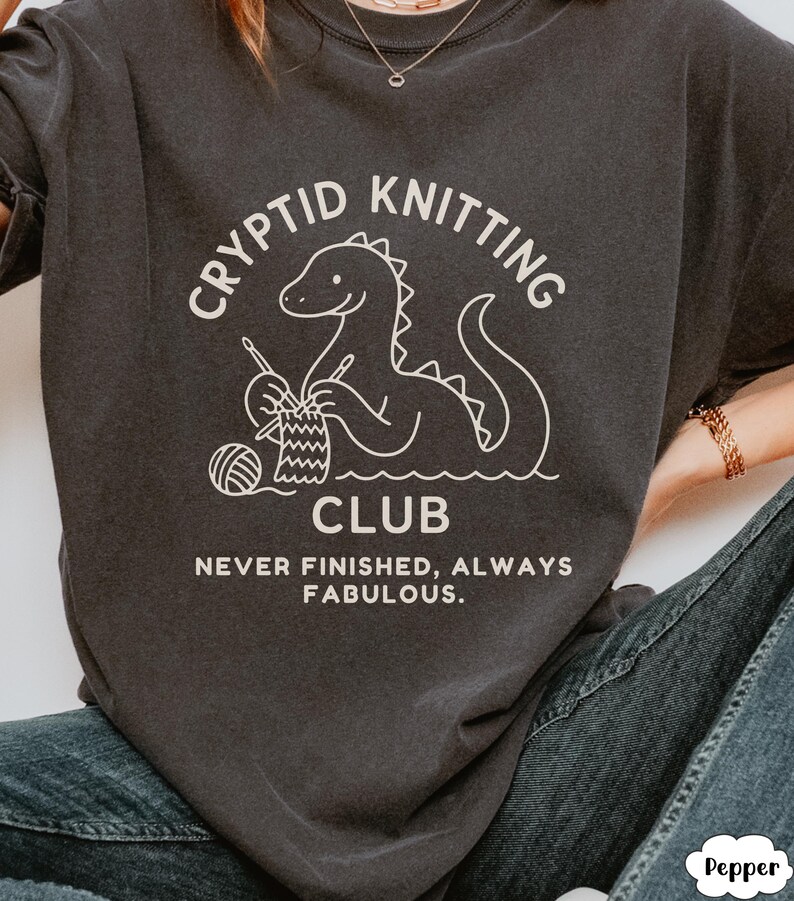 May include: Charcoal grey t-shirt featuring a white illustration of a dinosaur knitting. The text reads "Cryptid Knitting Club" above the image and "Never Finished, Always Fabulous" below. The graphic includes yarn and needles.