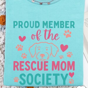 May include: A light blue t-shirt with the text "PROUD MEMBER of the RESCUE MOM SOCIETY" in teal and pink. The design includes paw prints, hearts, and a dog illustration. A delicate butterfly necklace is visible at the neckline.