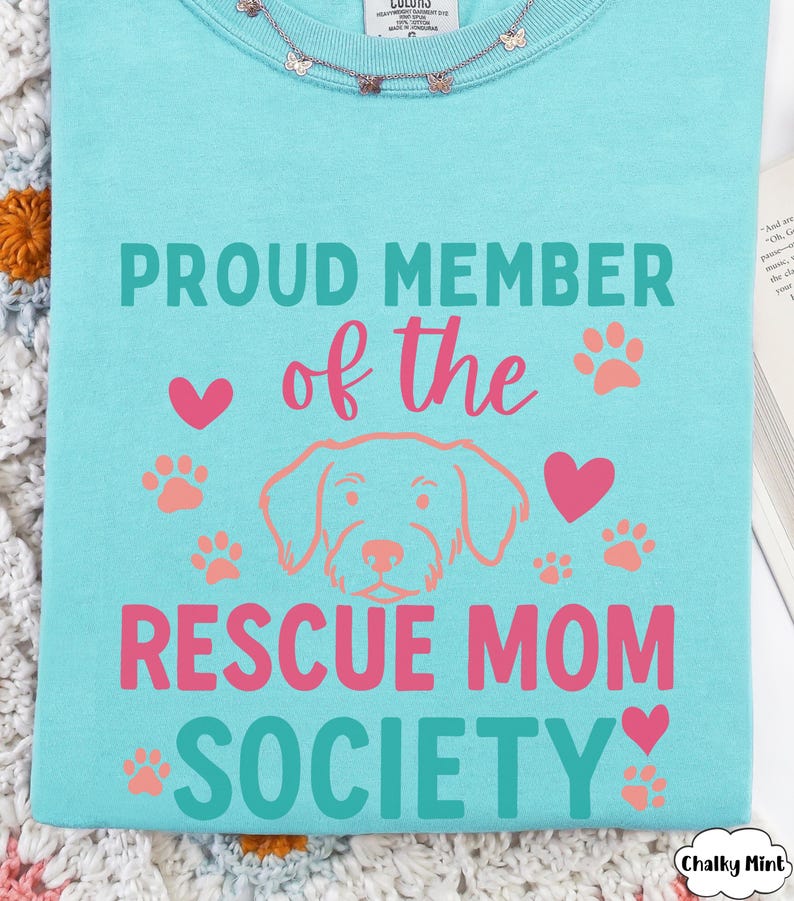 May include: A light blue t-shirt with the text "PROUD MEMBER of the RESCUE MOM SOCIETY" in teal and pink. The design includes paw prints, hearts, and a dog illustration. A delicate butterfly necklace is visible at the neckline.