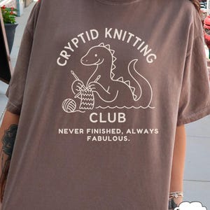 May include: Brown t-shirt featuring a white graphic of a cryptid knitting and the words "Cryptid Knitting Club". The text "Never Finished, Always Fabulous" is below the graphic. The shirt has short sleeves.
