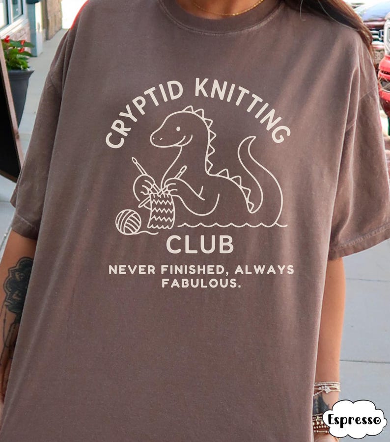 May include: Brown t-shirt featuring a white graphic of a cryptid knitting and the words "Cryptid Knitting Club". The text "Never Finished, Always Fabulous" is below the graphic. The shirt has short sleeves.