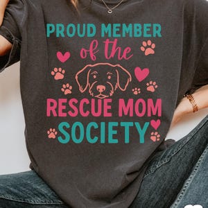 May include: Dark gray t-shirt with the text "PROUD MEMBER of the RESCUE MOM SOCIETY" in teal and pink. The design includes paw prints, hearts, and a dog illustration.