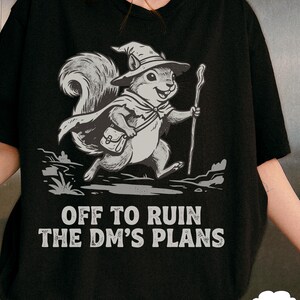 May include: Black t-shirt featuring a cartoon squirrel dressed as a wizard, holding a staff and wearing a hat and cape. The text "OFF TO RUIN THE DM'S PLANS" is printed below the squirrel illustration.