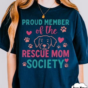 May include: Navy blue t-shirt with the text "PROUD MEMBER of the RESCUE MOM SOCIETY" in teal and pink. The design includes paw prints, hearts, and a dog outline. The shirt has a relaxed fit.