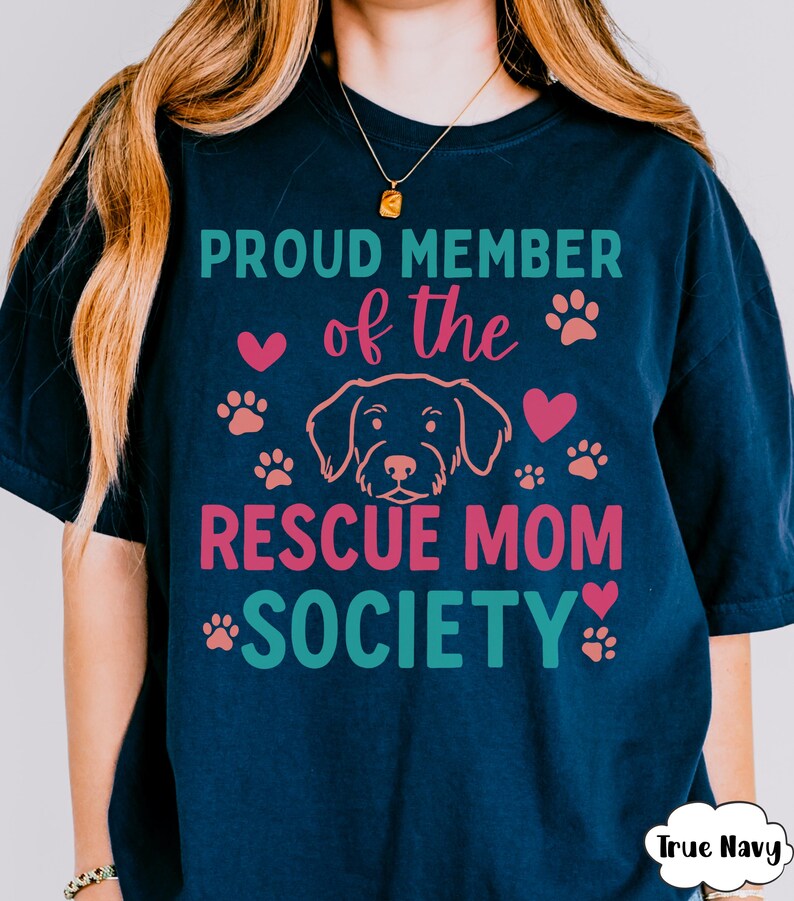 May include: Navy blue t-shirt with the text "PROUD MEMBER of the RESCUE MOM SOCIETY" in teal and pink. The design includes paw prints, hearts, and a dog outline. The shirt has a relaxed fit.