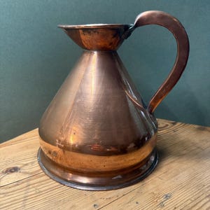 May include: A polished copper pitcher with a wide, conical base that tapers toward a flared rim. The vessel features a single curved handle attached to the side, showcasing a warm, metallic finish with subtle surface variations and reflections.