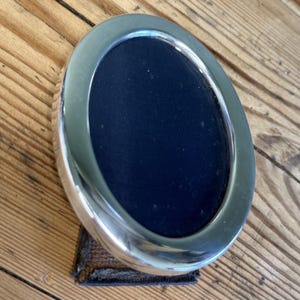 Op de afbeelding: Ovale zilverkleurige fotolijst met een donkerblauwe binnenkant. De lijst heeft een gepolijste afwerking en staat op een kleine, rechthoekige, donkergekleurde basis. De lijst wordt op een houten oppervlak gepresenteerd.