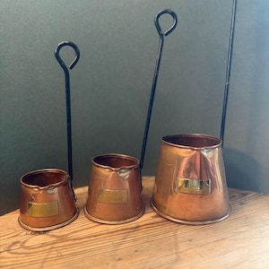 May include: A set of three graduated copper measuring cups with long black metal handles and circular loops at the ends. Each cup features a small pouring spout and a rectangular brass plaque attached to the front of the polished copper body.