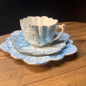 Op de afbeelding: Een theekop en schotel set van porselein met een geschulpte rand en gouden afwerking. Het witte keramiek is versierd met een lichtblauw bloemenpatroon. De kop heeft een gebogen handvat en een gegroefde vorm, passend bij de schotel eronder.