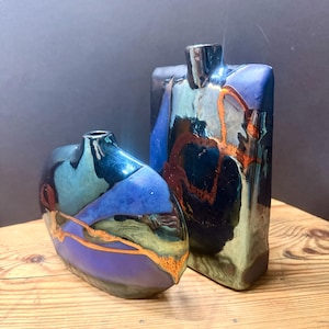 May include: Two ceramic vases with a glossy, multi-colored glaze featuring deep blue, teal, and metallic copper accents. The shorter vase has an oval shape, while the taller vase is rectangular with rounded edges, both featuring narrow necks.