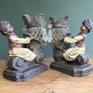 Può includere: Un paio di fermalibri decorativi con figure di persone con turbanti, sedute su basi ornate. Le figure indossano gilet e turbanti marroni, con camicie e pantaloni color crema. I fermalibri hanno una combinazione di colori marrone scuro e verde.