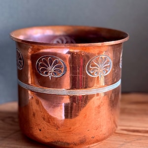 May include: A cylindrical copper pot with a polished metallic finish. The upper section features a repeating embossed pattern of stylised floral motifs within oval frames, separated from the lower section by a thin, horizontal silver-toned band.