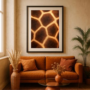 Op de afbeelding: Een ingelijste print met een close-up van het vachtpatroon van een giraffe. Het kunstwerk toont een herhalend patroon van bruine en bruingeel vormen, omlijst in zwart. De print hangt boven een oranje bank met decoratieve kussens en een salontafel met glazen blad.