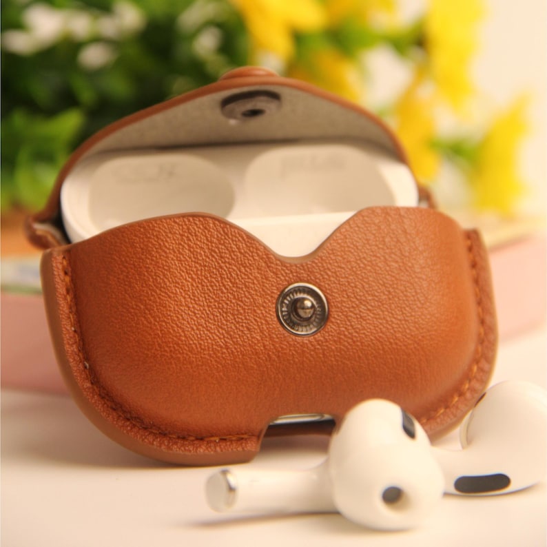 May include: A brown leather earbud case with a snap closure, open to reveal white earbuds inside. Two white earbuds are resting next to the case. The case has a stitched border and a silver snap.