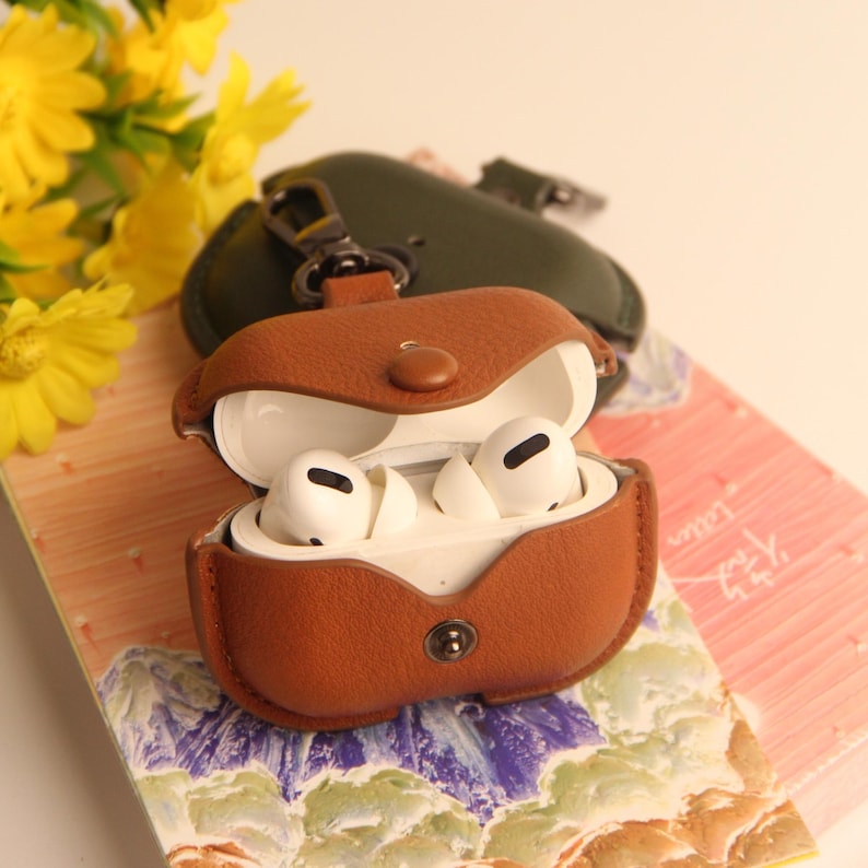 May include: A brown leather AirPods Pro case with a snap closure, open to reveal white earbuds. A green leather case and yellow flowers are in the background. The case has a silver clasp.