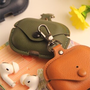 May include: Close-up of two leather AirPod cases in olive green and tan, with silver carabiner clips. White wireless earbuds are visible in the lower left corner. The cases have a button closure and are designed to protect the earbuds.