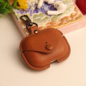 May include: A brown leather AirPod case with a snap closure and a metal clasp. The case is designed to protect the AirPods and has a smooth, textured surface. The case is shown on a decorative surface with white earbuds.