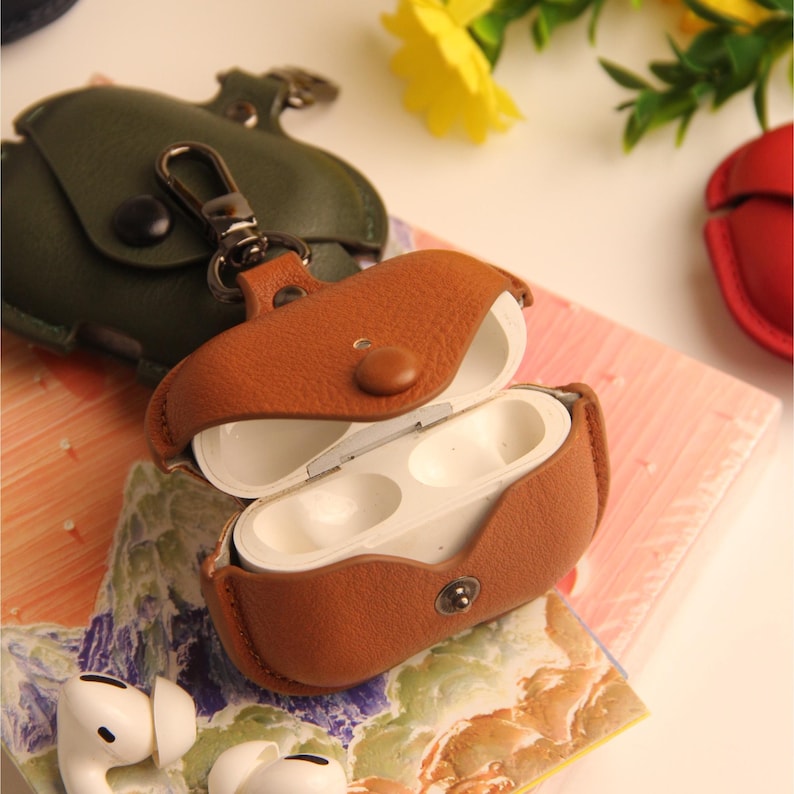 May include: A brown leather AirPod case with a snap closure, open to reveal white earbuds. A green leather case and red case are visible in the background. The case has a silver clasp for attaching to a bag or keychain.