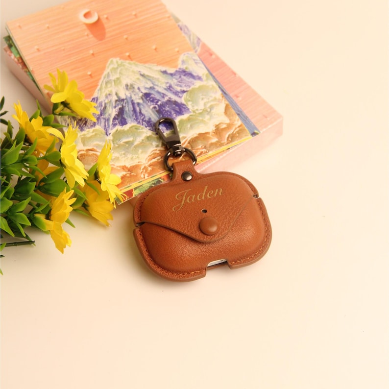 May include: A brown leather AirPod case with the name "Jaden" engraved on the front. The case has a snap closure and a metal clasp. The case is next to a colorful notebook and yellow flowers.