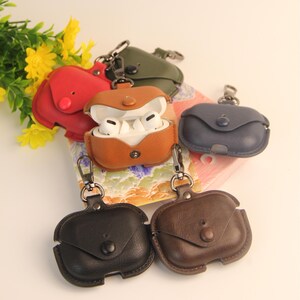 May include: A collection of leather AirPod cases in various colors, including red, brown, green, blue, black, and brown. Each case has a snap closure and a metal clasp for attaching to a keychain or bag. The cases are designed to protect and carry wireless earbuds.