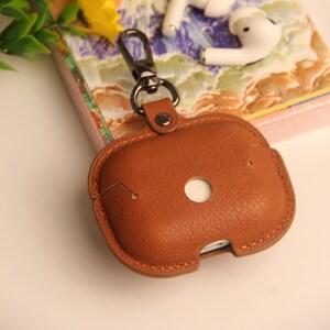 May include: A brown leather AirPods case with a metal clasp and a key ring. The case has a circular opening and a stitched border. White wireless earbuds rest on a patterned book in the background.